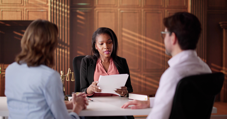 A female lawyer sits at a desk reviewing documents while speaking with a man and woman seated across from her. The setting has warm lighting and a wood-paneled background, with legal books and scales visible on the desk.