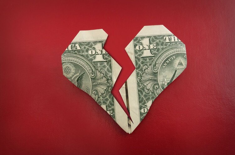 A one-dollar bill is folded into the shape of a broken heart on a red background. The design symbolizes financial separation or the economic impact of a breakup.