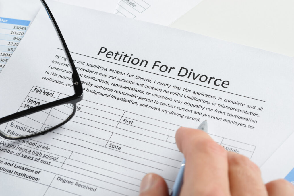 Close-up of a hand holding a pen over a “Petition for Divorce” form, with eyeglasses resting on the paper. does it matter who files for divorce first
