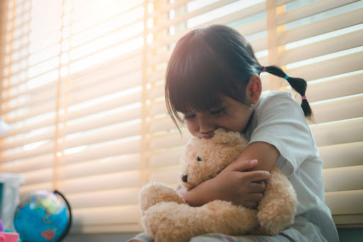 Young girl hugging a plush teddy bear, wearing a white shirt, sitting by window blinds with soft sunlight filtering through. what can be used against you in a custody battle